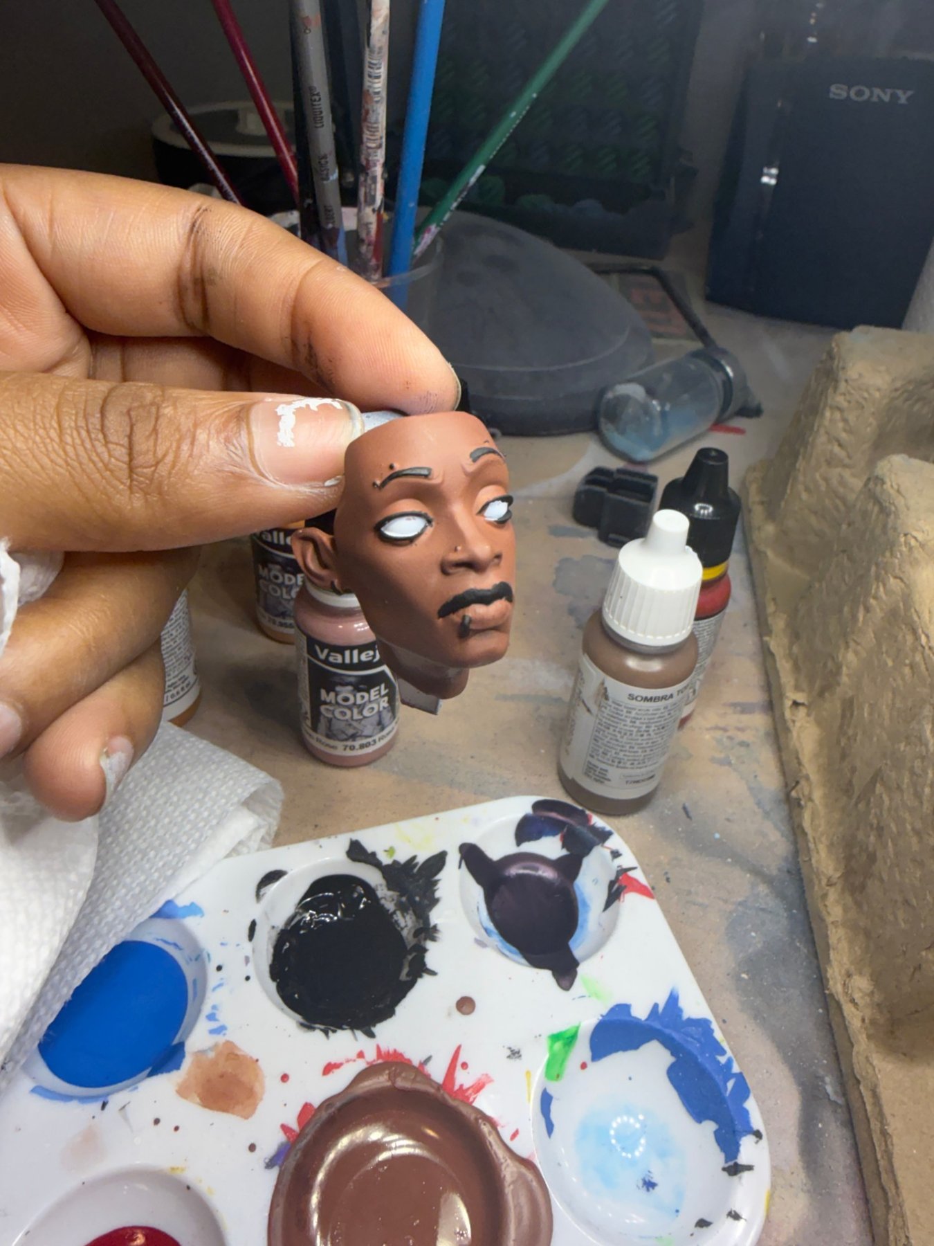 A hand-painted miniature head, mid-progress, held between two fingers — base skin tone and inked features, paint bottles and a mixing palette in the background.
