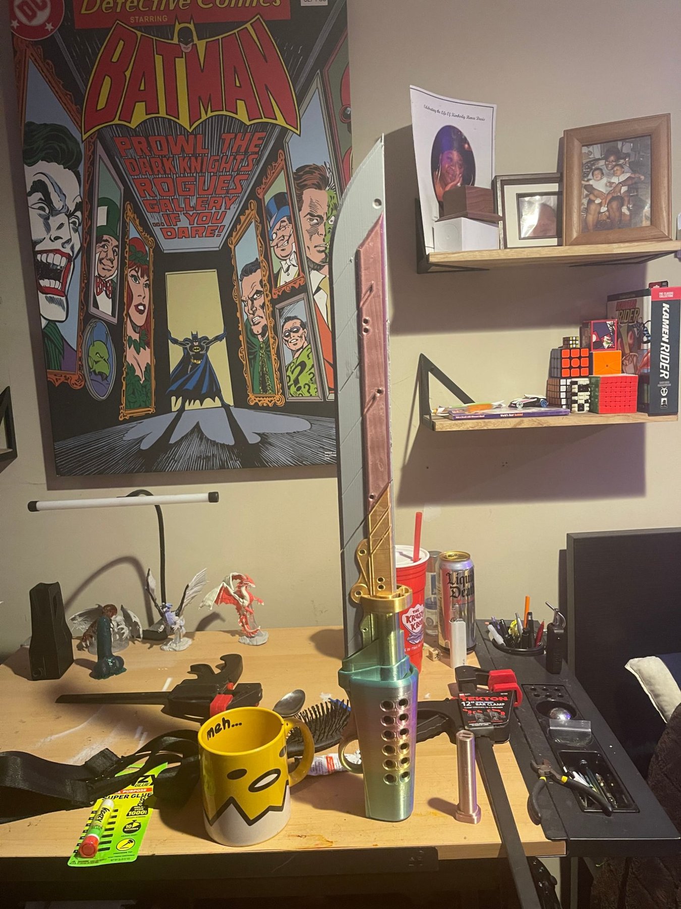 An unfinished 3D printed prop blade standing on a desk, unpainted sections still bare plastic, next to a comic poster and desk clutter.
