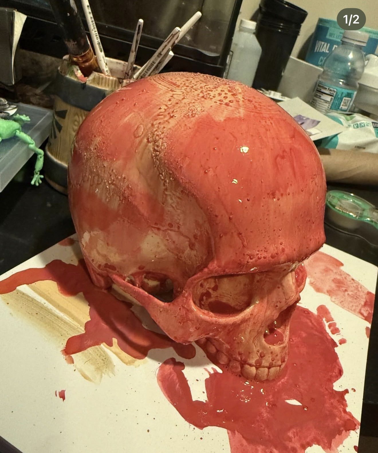 A life-size printed skull washed in wet red paint, pooling onto white paper on a cluttered desk.