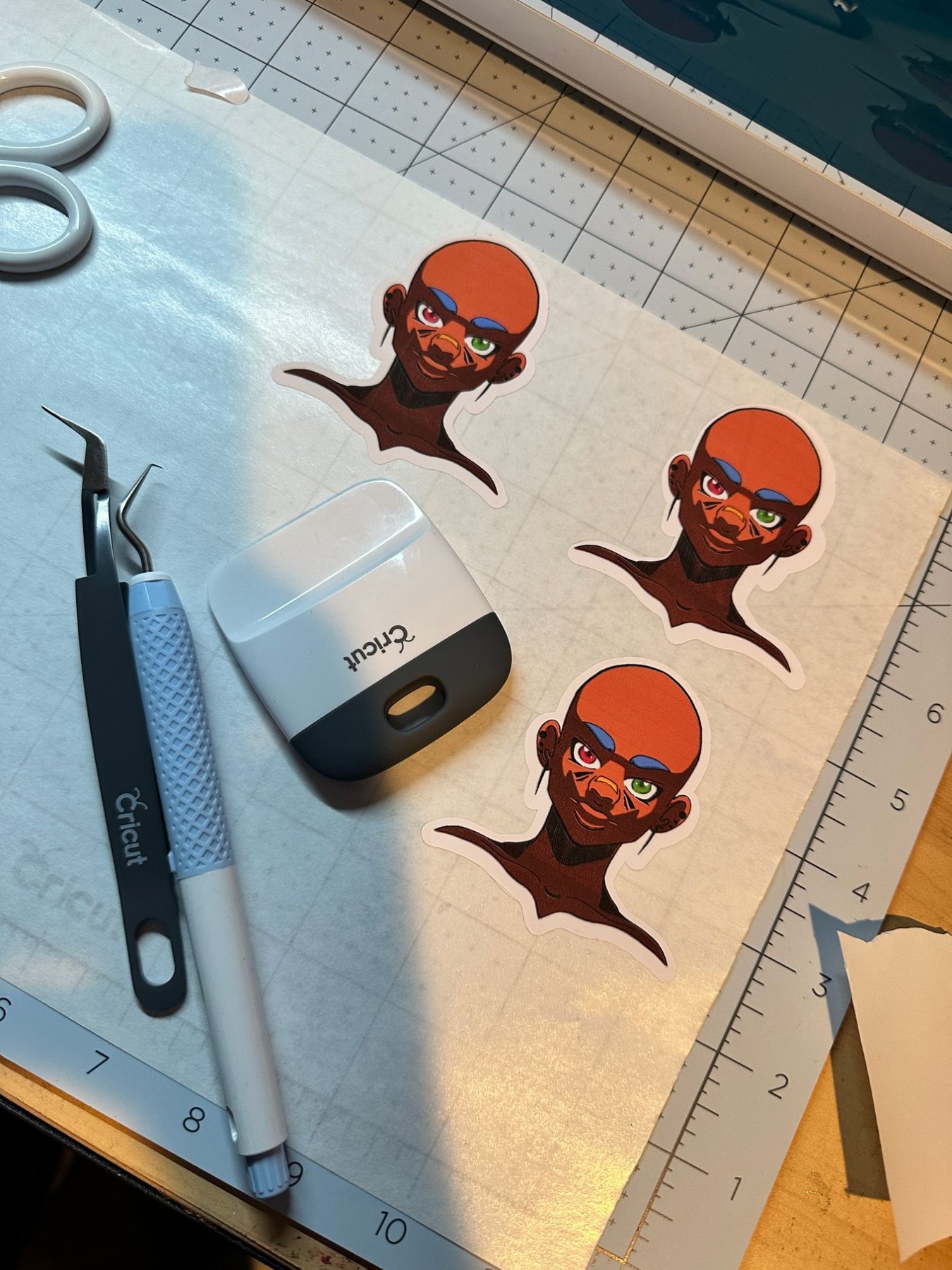 Three illustrated character stickers on a cutting mat next to Cricut weeding tools and a scraper.